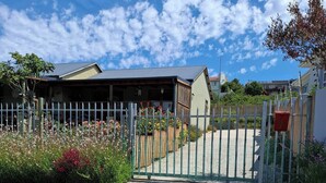A gravel driveway leading to a house with a white fence, a wooden gate, and a red mailbox. - Petite Cottage Plett Close to Town (Plettenberg Bay)
