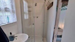 A bathroom with a glass shower, a sink, and a window with a decorative curtain. - Petite Cottage Plett Close to Town (Plettenberg Bay)