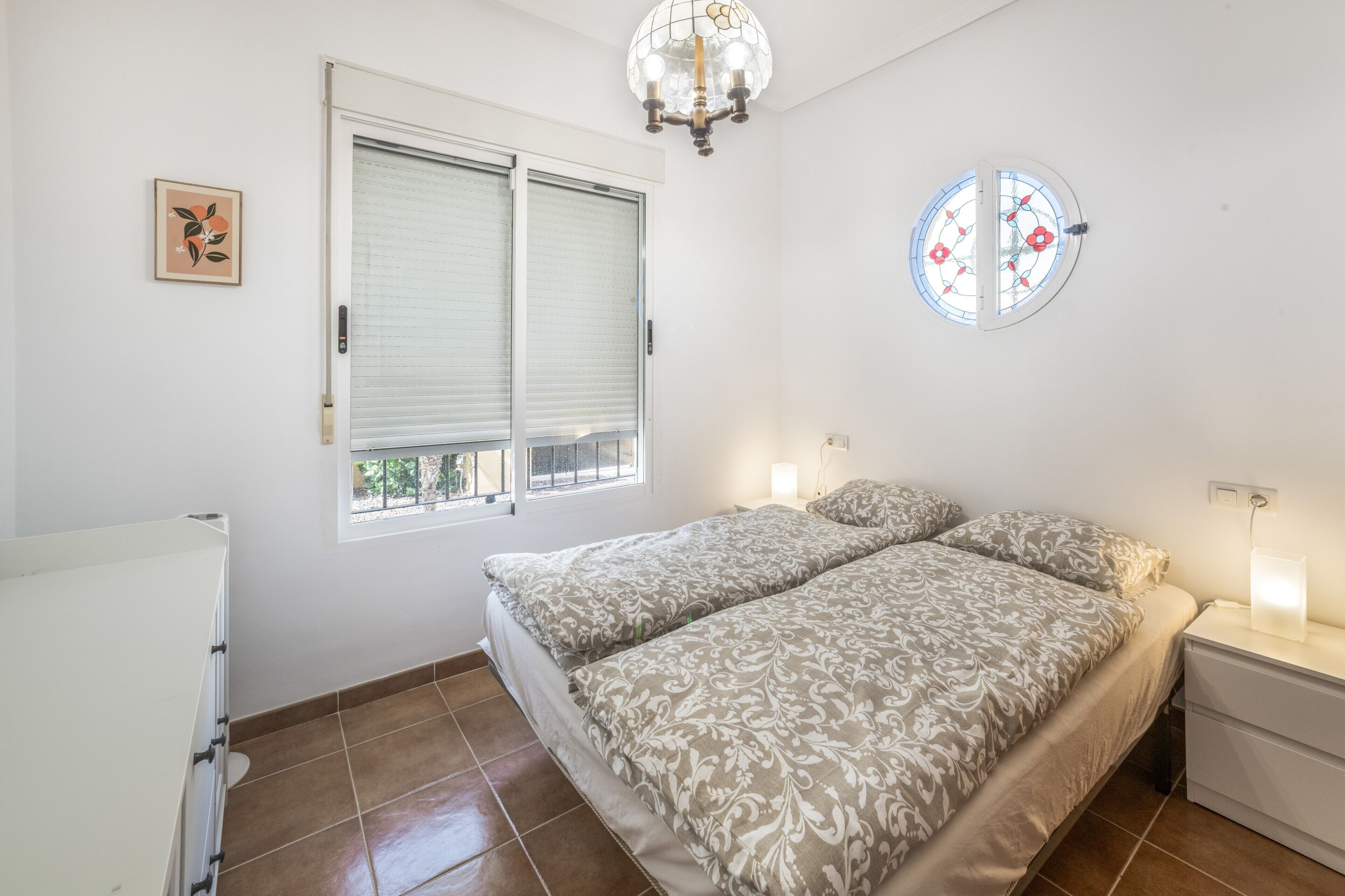 Une chambre à coucher comprenant un lit, des tables de chevet, une fenêtre avec des stores, un lustre et un luminaire fixé au mur.