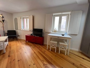 Una habitación con piso de madera, un escritorio blanco con silla, un aparador rojo, un espejo y una ventana con persianas.