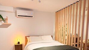 A bedroom with a bed, bedside table, wall-mounted air conditioner, and a window with vertical blinds.