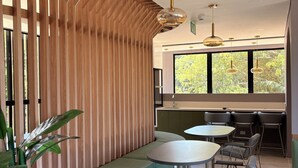 A modern interior with a curved wooden partition, a green cushioned bench, a round table with chairs, and large windows with a view of greenery.