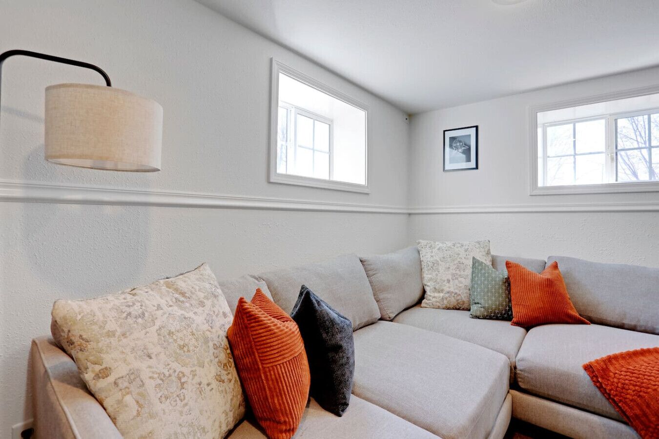 A living room with a sectional sofa, two windows, a lamp, and a framed picture on the wall.