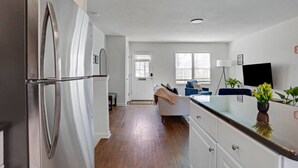 Una cocina moderna con un refrigerador de acero inoxidable, piso de madera, una zona de comedor con un sofá azul y un televisor de pantalla plana montado en la pared.