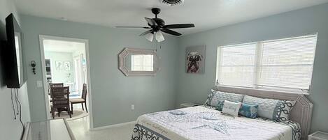 A bedroom with a bed, a ceiling fan, a window with blinds, a mirror, and a television on the wall.