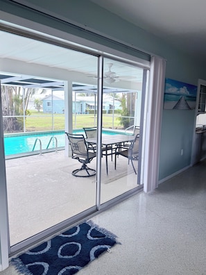 A room with a sliding glass door leading to a pool area, a dining table with chairs, and a painting on the wall.