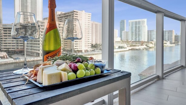 Mesa com garrafa de champanhe, duas taças e variedade de queijos e uvas numa varanda com vista para a cidade e para o mar.