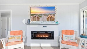 Ein Wohnzimmer mit Kamin, zwei Korbsessel mit Kissen, einem über dem Kamin montierten Fernseher und Blick auf einen Strand durch das Fenster.