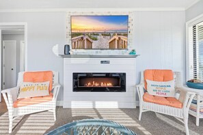 A living room with a fireplace, two wicker chairs with cushions, a television mounted above the fireplace, and a view of a beach through the window. - Golden Mile Oceanview Retreat (Myrtle Beach)