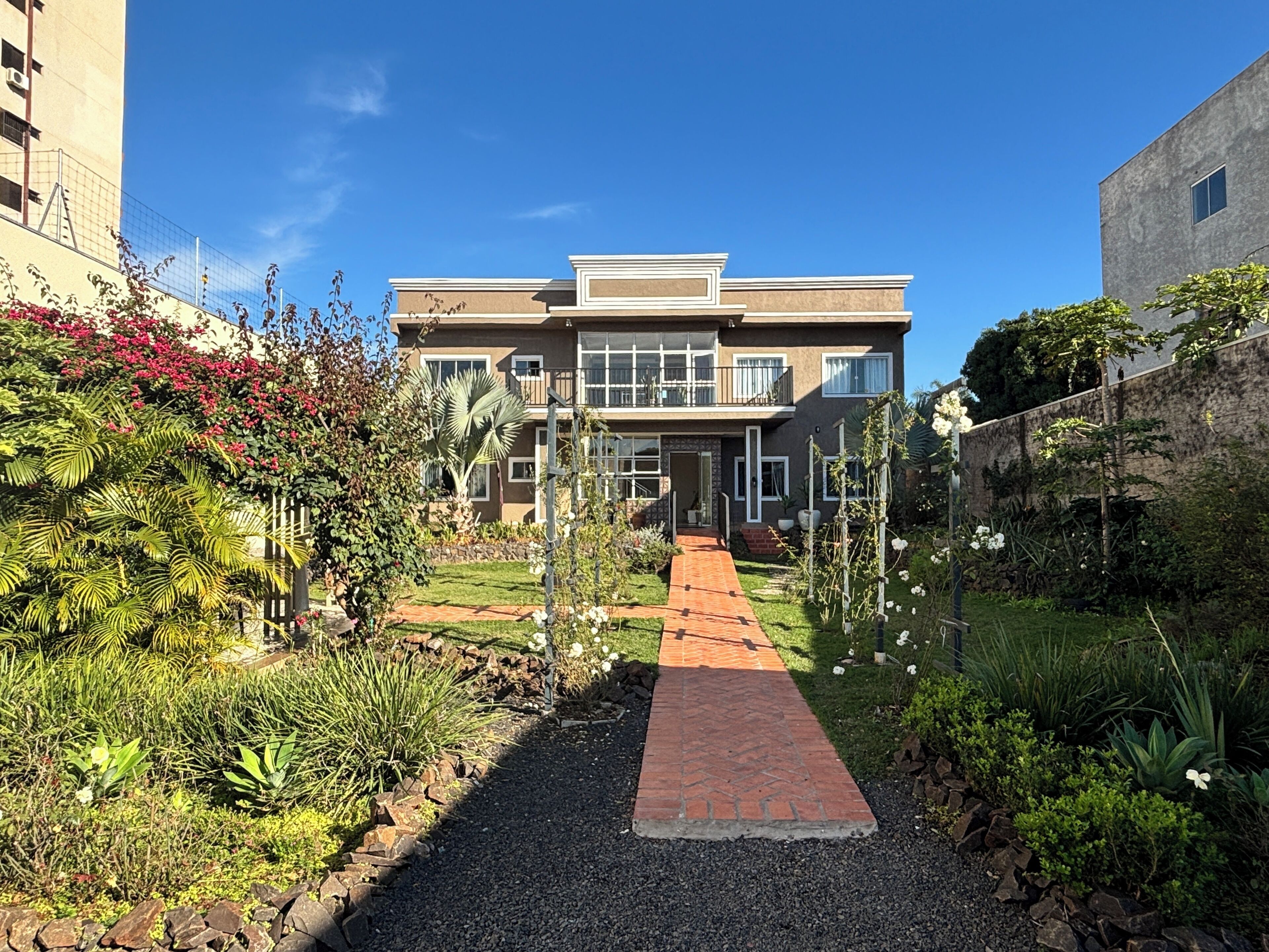 Uma casa moderna de dois andares com um caminho pavimentado que leva à entrada, cercada por vegetação exuberante e plantas floridas.