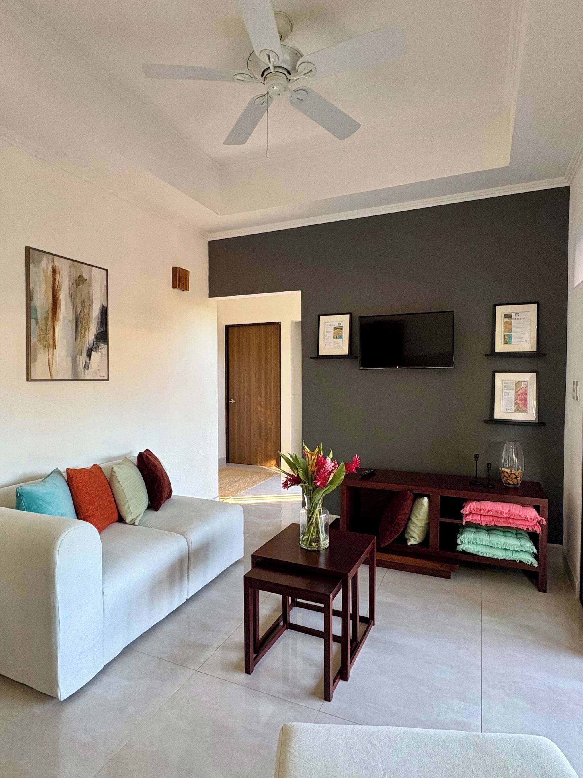 Una sala de estar moderna con un sofá blanco, una mesa de centro, un televisor montado en la pared y un ventilador de techo.