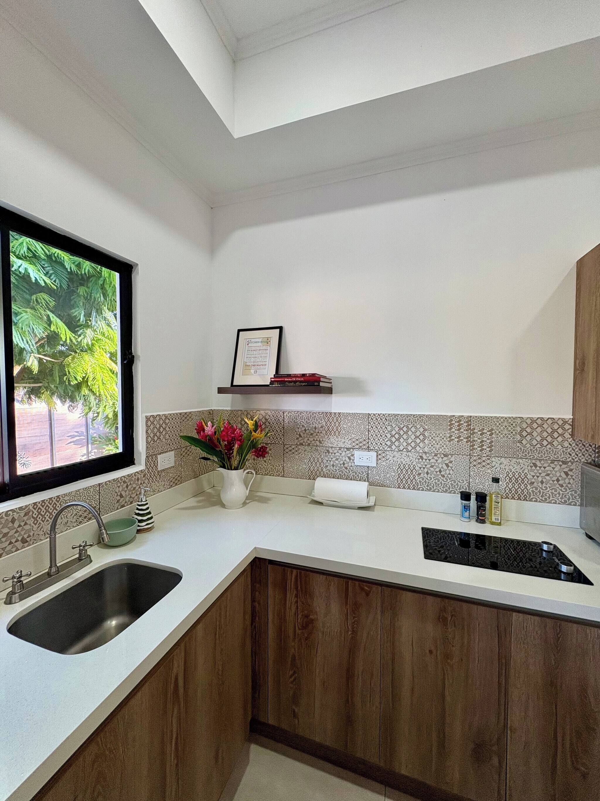 Cocina moderna con pileta integrada, armarios de madera, ventana con vista a vegetación, salpicadero de azulejos y un estante pequeño con libros y una foto enmarcada.