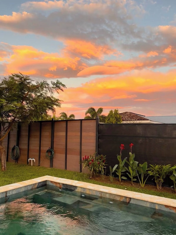 A swimming pool with a wooden fence and plants, under a sunset sky.