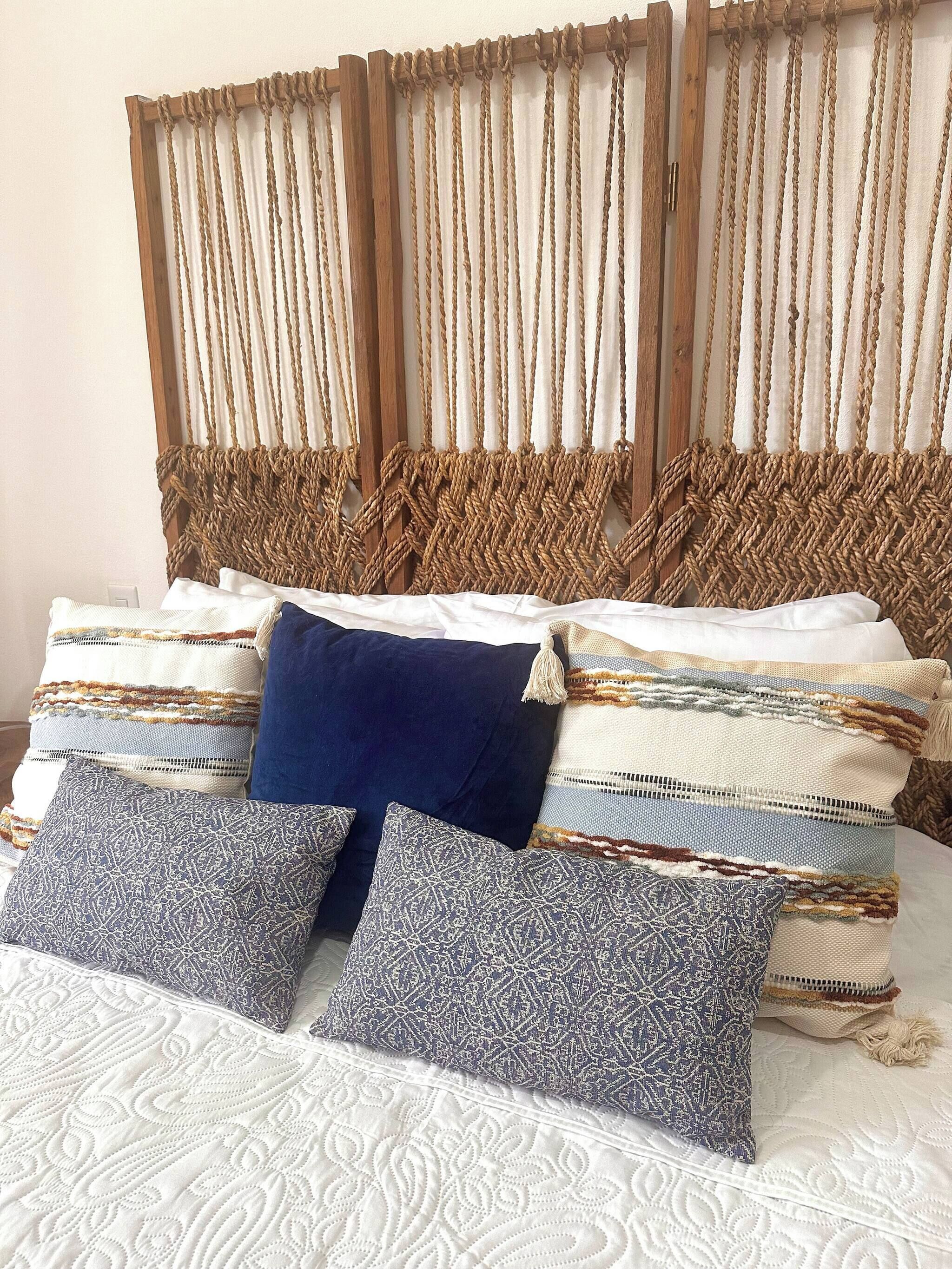 Una cama con cabecera de macramé, varios cojines estampados y una colcha acolchada blanca.