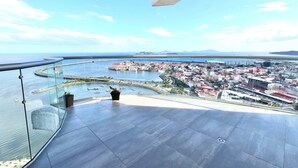 Un balcon doté d’une balustrade en verre, offrant une vue sur un paysage urbain côtier avec une baie, des bâtiments et un pont.