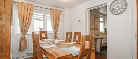 Un comedor con una mesa y sillas de madera, cortinas estampadas, un reloj en la pared y vistas a la cocina.