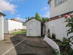 Een smalle steeg met een witte schuur en een huis aan de ene kant, en een witte muur met een tuin aan de andere kant.