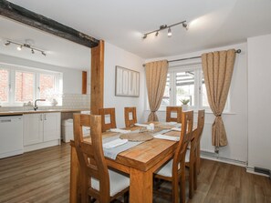 A dining area with a wooden table and chairs, a kitchen with white cabinets, and patterned curtains. - Corner House (Shrewsbury)