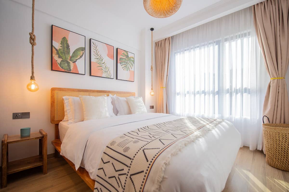 A bedroom with a bed, a bedside table, a hanging light fixture, and a window with curtains.