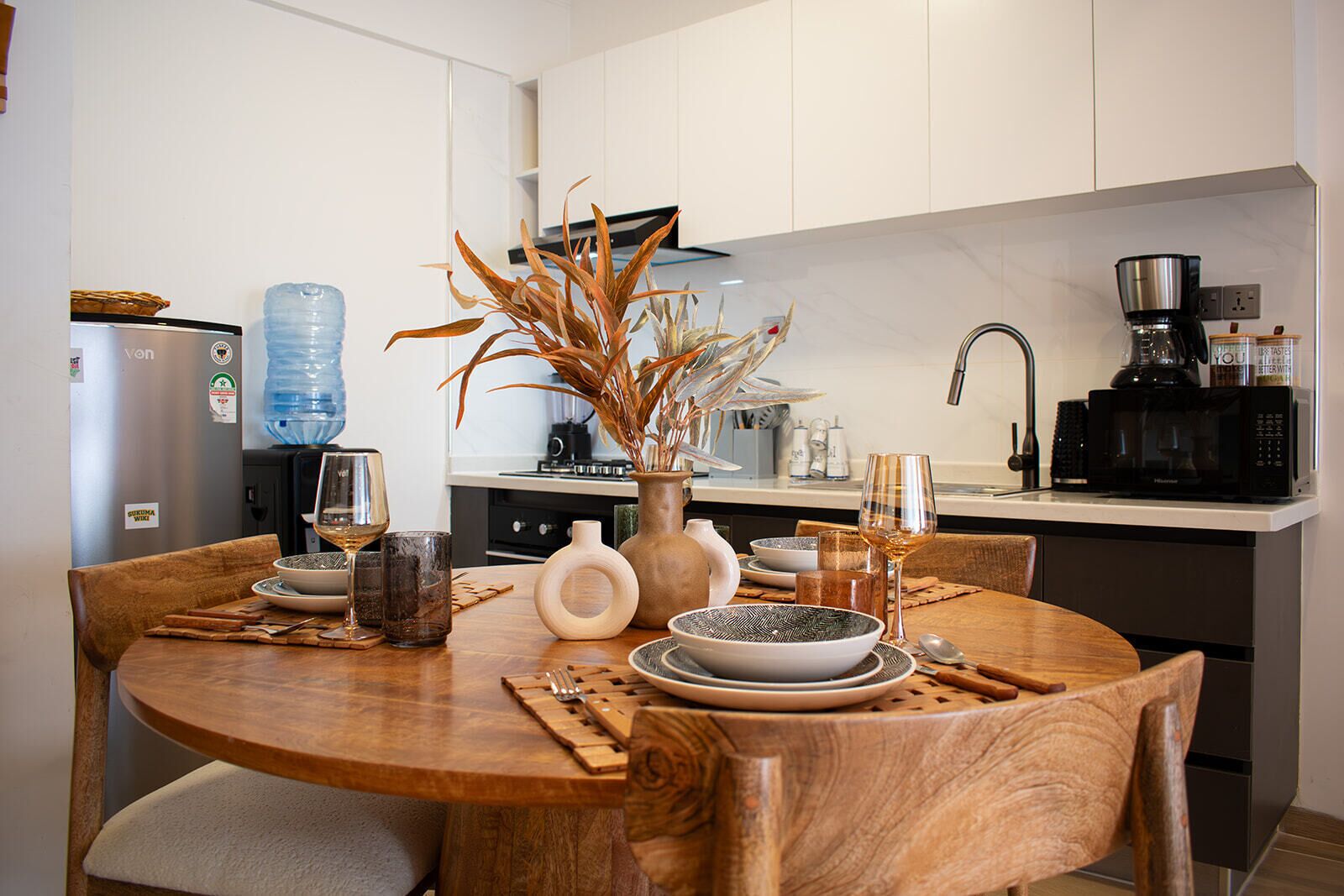 A modern kitchen with a dining table set for two, featuring a water dispenser, a coffee maker, and a microwave.
