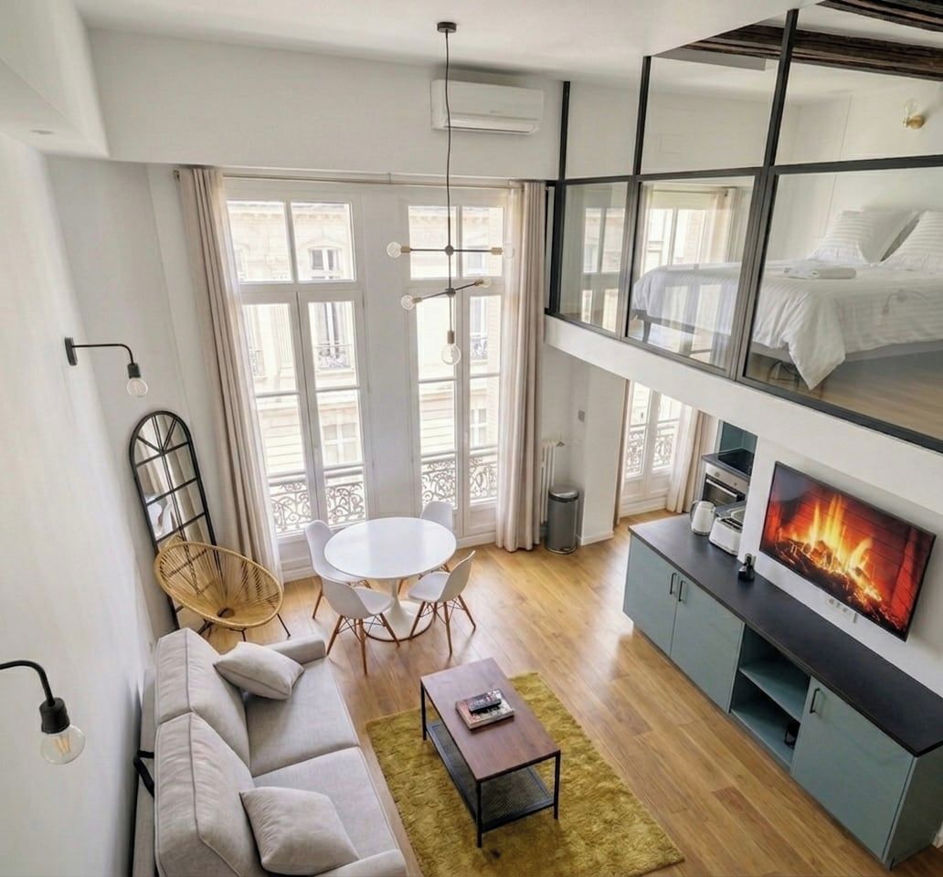 A modern living room with a sofa, a round dining table, a coffee table, a TV with a fireplace display, and a balcony with a railing.
