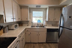 A compact kitchen with white cabinets, a stainless steel refrigerator, a dishwasher, a toaster, and a window with a view of the outside.