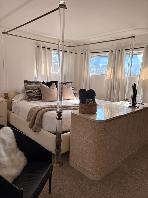A bedroom with a four-poster bed, a bedside table, a chair, and a view of the outdoors.