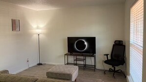 A living room with a TV, a floor lamp, a black office chair, and a window with blinds. - 1x1 Apt Right by Pease Park (Austin)