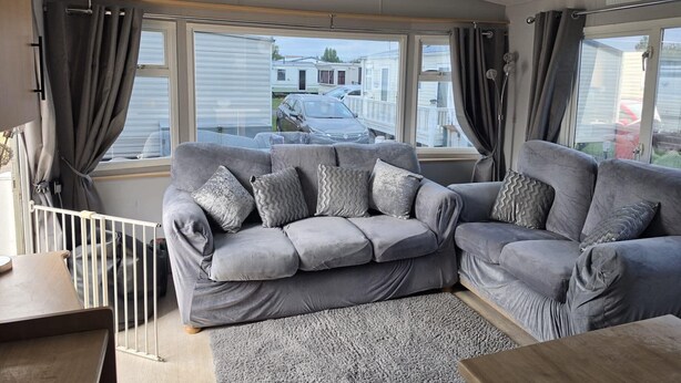 A modern living room with a grey sofa, a wooden coffee table, and a view of a residential area through large windows. - 158 South Happy Days - Sleeps 8 (Skegness)