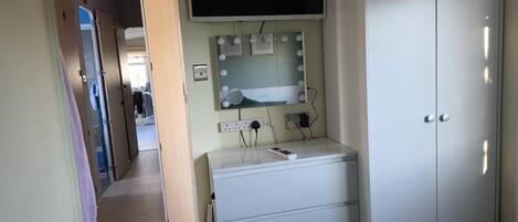 A bedroom with a bed, a dresser, a vanity with a mirror and lights, and a TV on the wall.