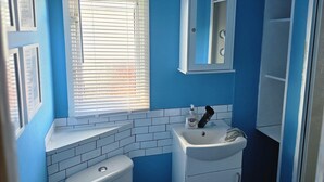 A bathroom with a blue wall, white tiles, a sink, a toilet, and a window with blinds. - 158 South Happy Days - Sleeps 8 (Skegness)