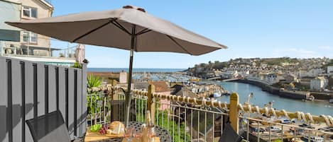 A balcony with a table, chairs, and an umbrella overlooking a harbor with boats and buildings.