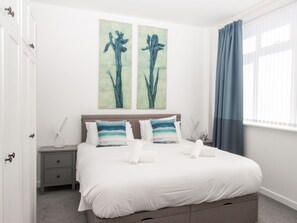 Un dormitorio con cama, dos almohadas, una mesita de noche, un cuadro en la pared y una ventana con cortinas.