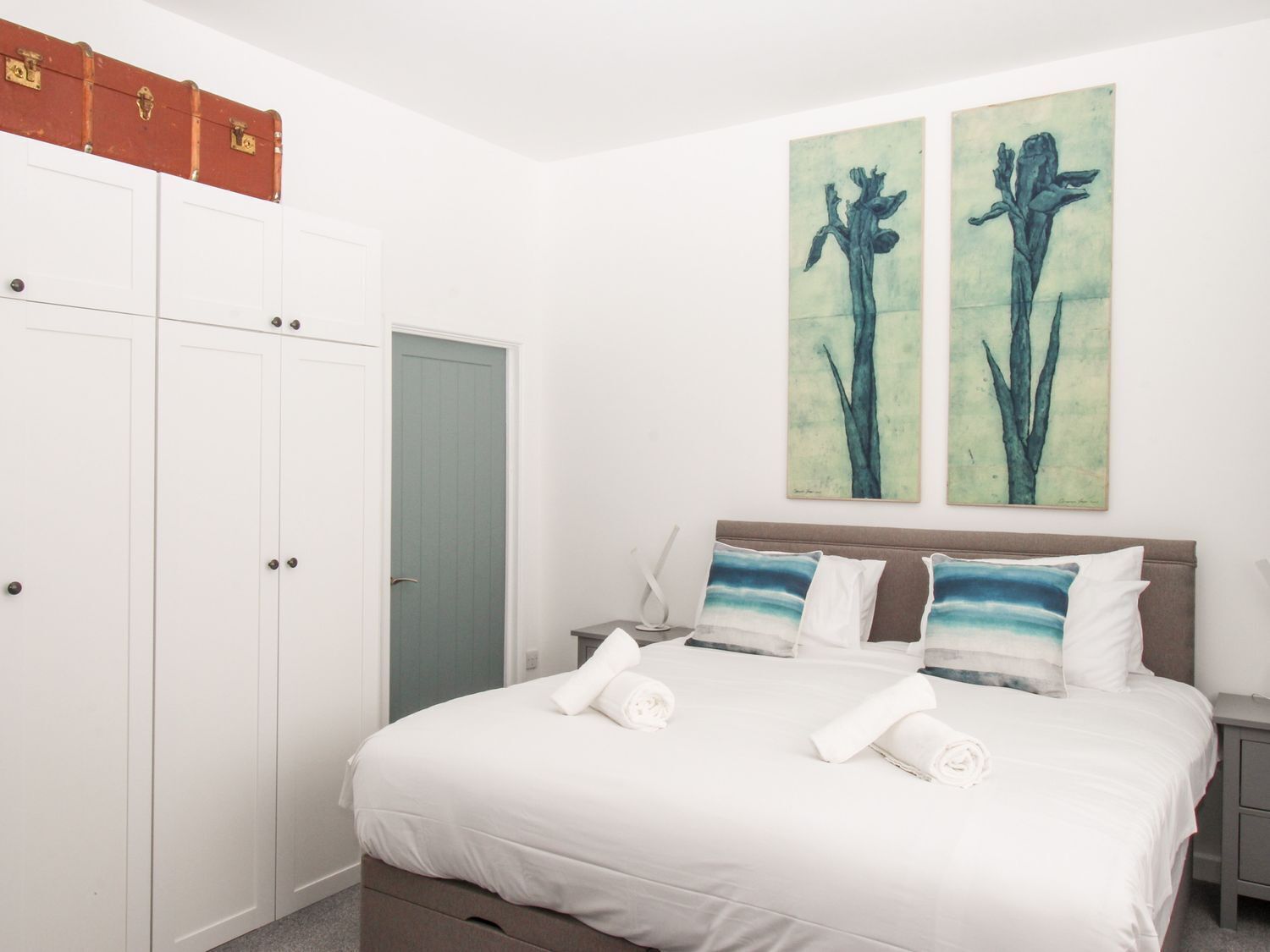 A bedroom with a bed, two paintings, a wardrobe, and a suitcase on top.