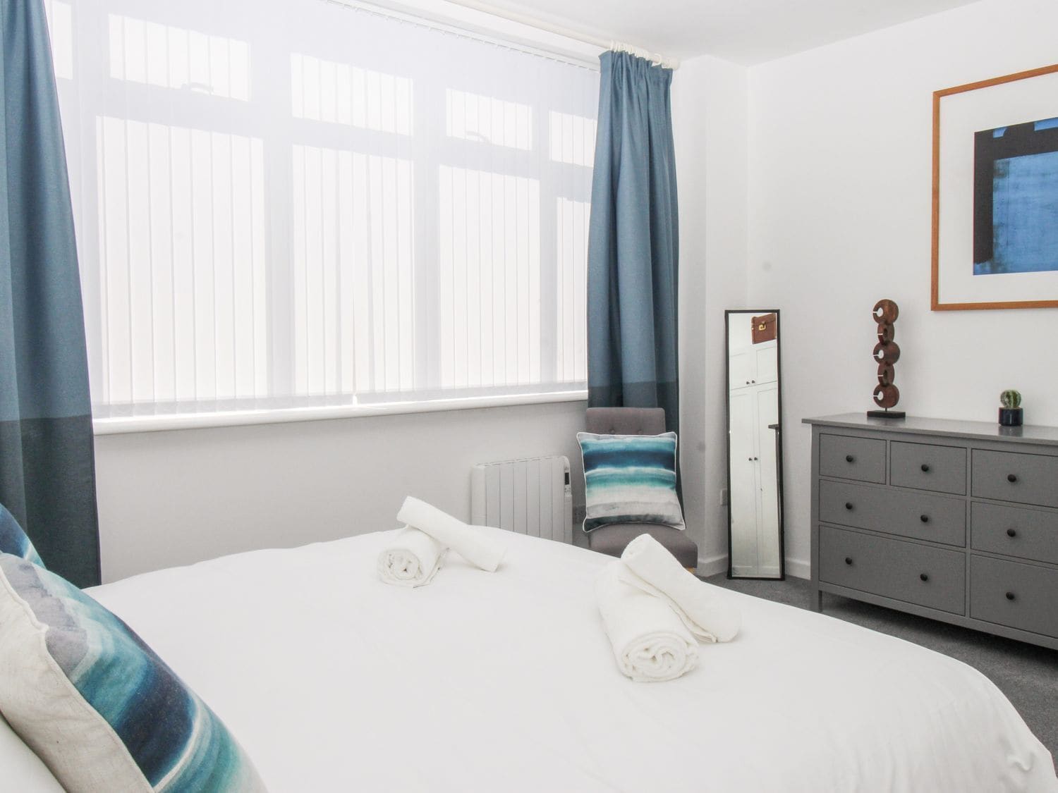 A hotel room with a bed, a dresser, a radiator, a window with curtains, and a framed picture on the wall.