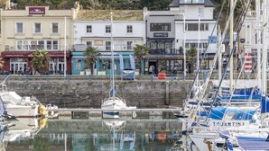 Un port de plaisance avec des bateaux amarrés, un bâtiment situé au bord de l’eau abritant des boutiques et un mur de pierre.