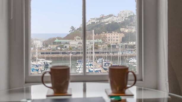 Une vue à travers une fenêtre, avec deux tasses posées sur une table, donnant sur une marina où l’on aperçoit des bateaux et des bâtiments sur une colline.