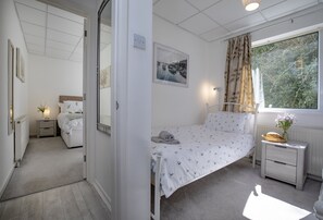 A bedroom with a single bed, a bedside table, a nightstand, a window with curtains, a radiator, and a wall-mounted light fixture. - Enjoy a relaxing seaside escape at Ocean Side Apartment (Brixham)