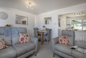 A living room with two grey sofas, a coffee table, and a dining table with chairs. There are two wall clocks and a ceiling light fixture. - Enjoy a relaxing seaside escape at Ocean Side Apartment (Brixham)