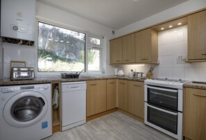 A modern kitchen with wooden cabinets, a built-in oven, a dishwasher, and a window with a view of trees. - Enjoy a relaxing seaside escape at Ocean Side Apartment (Brixham)