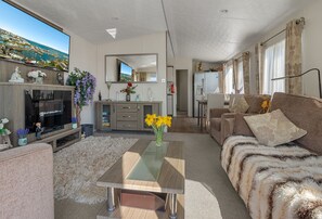A modern living room with a flat-screen TV, a glass-top coffee table, a sofa, and a kitchen area in the background.