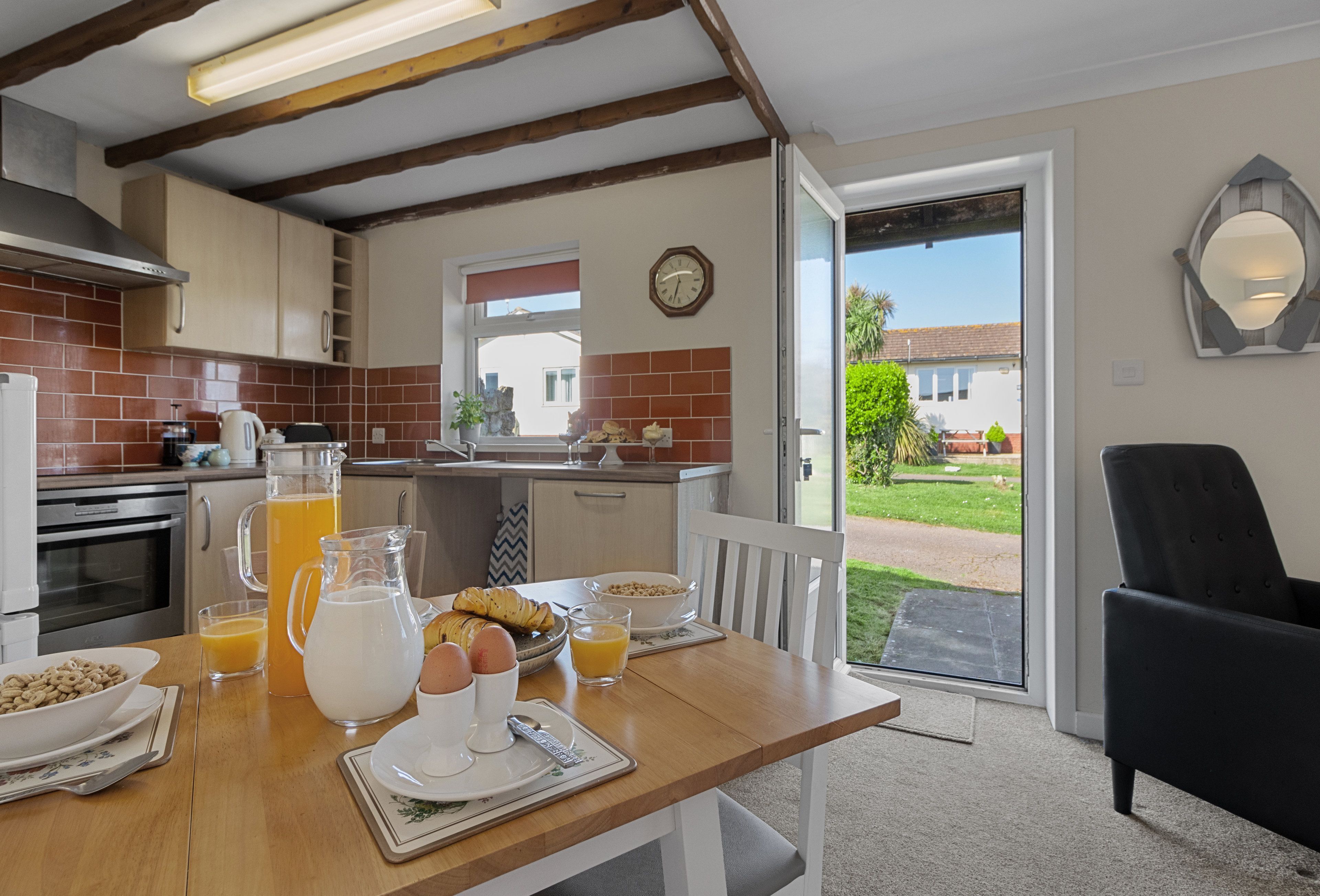Ein Frühstückstisch mit einer Karaffe, Gläsern und Croissants, eine Küche mit Herd und Backofen sowie der Blick auf einen Garten durch eine Schiebetür.