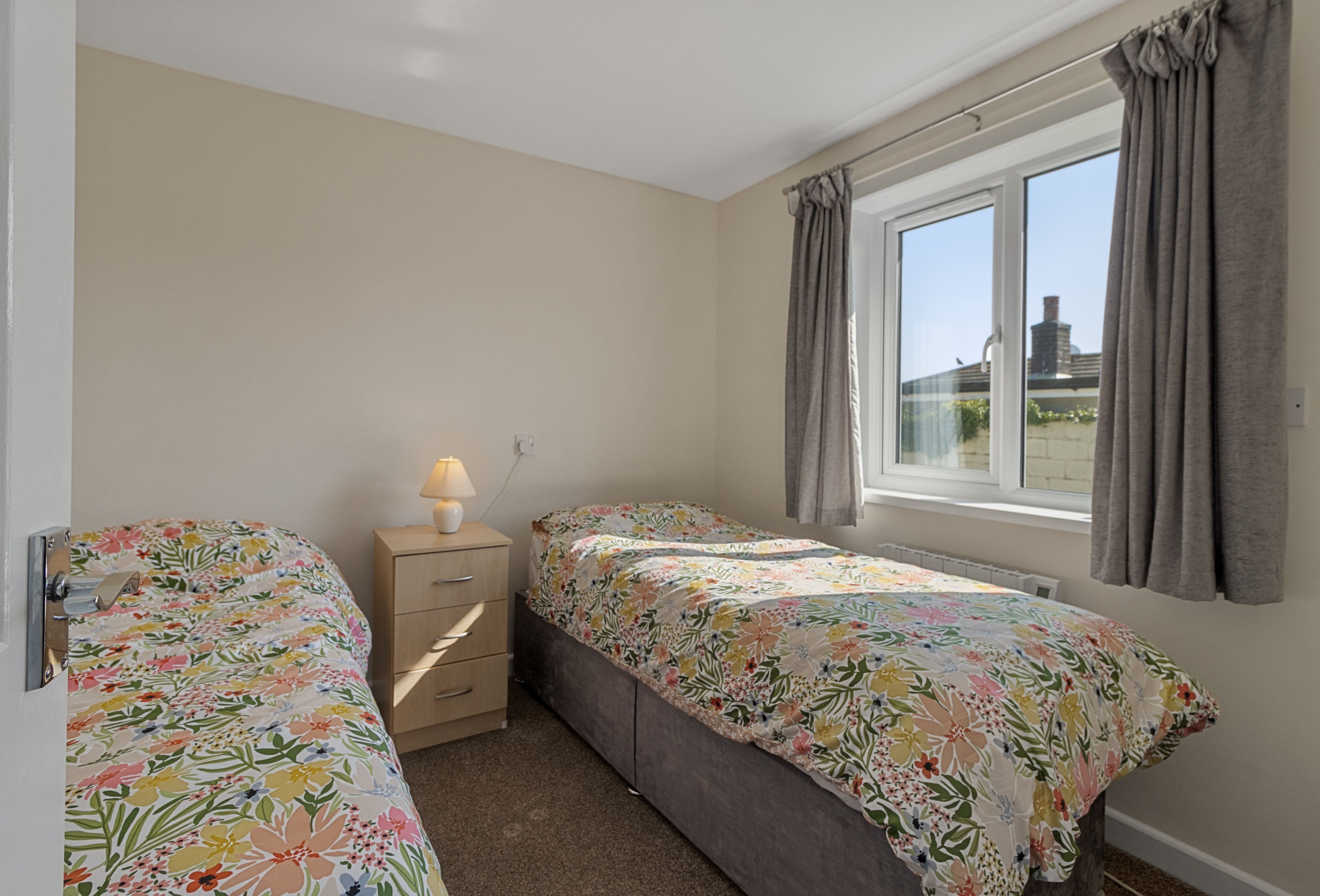 A bedroom with two single beds, a bedside table with a lamp, a window with curtains, and a view of the outside.
