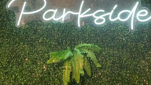 En neonskylt med texten ”Rease Parkside” mot en bakgrund av grönt lövverk.