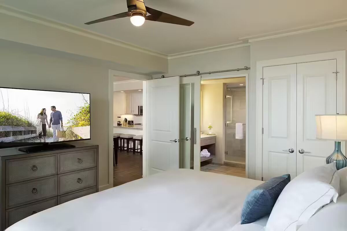Une chambre d’hôtel équipée d’un lit, d’une télévision, d’un ventilateur de plafond, d’une salle de bain avec un miroir et d’une porte donnant sur un coin cuisine.