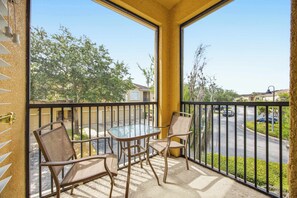 A balcony with a glass table and chairs, a view of a street and houses, and a yellow wall. - Cozy Disney Getaway 2BR  5 Mins to Parks (Kissimmee)