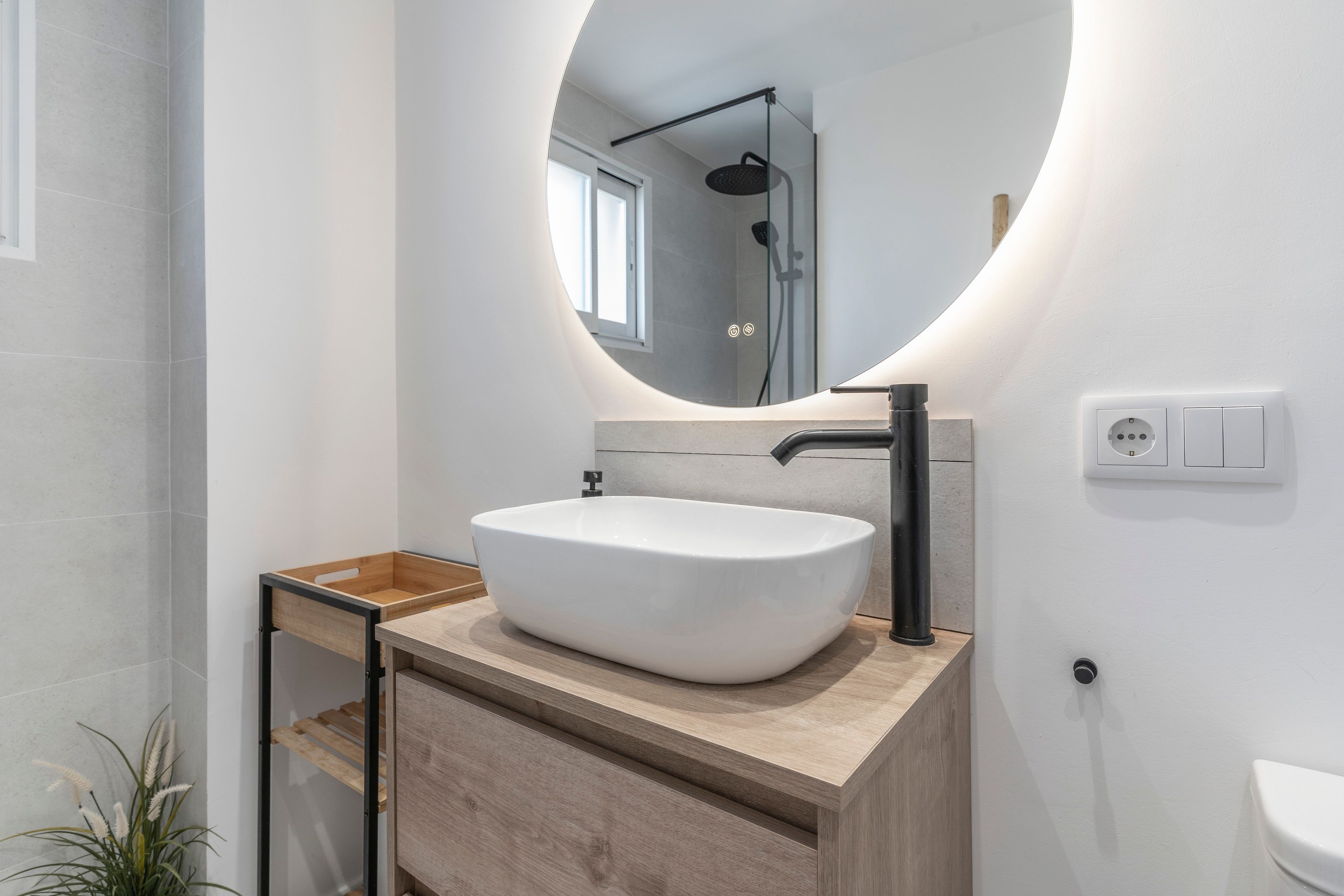 A modern bathroom with a white sink, a wooden vanity, a round mirror, and a shower visible in the background.