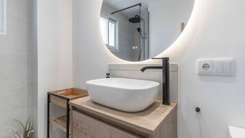 A modern bathroom with a white sink, a wooden vanity, a round mirror, and a shower visible in the background.