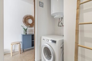 Une buanderie équipée d’un lave-linge, d’un petit tabouret, d’un miroir décoratif et d’un porte-serviettes fixé au mur.