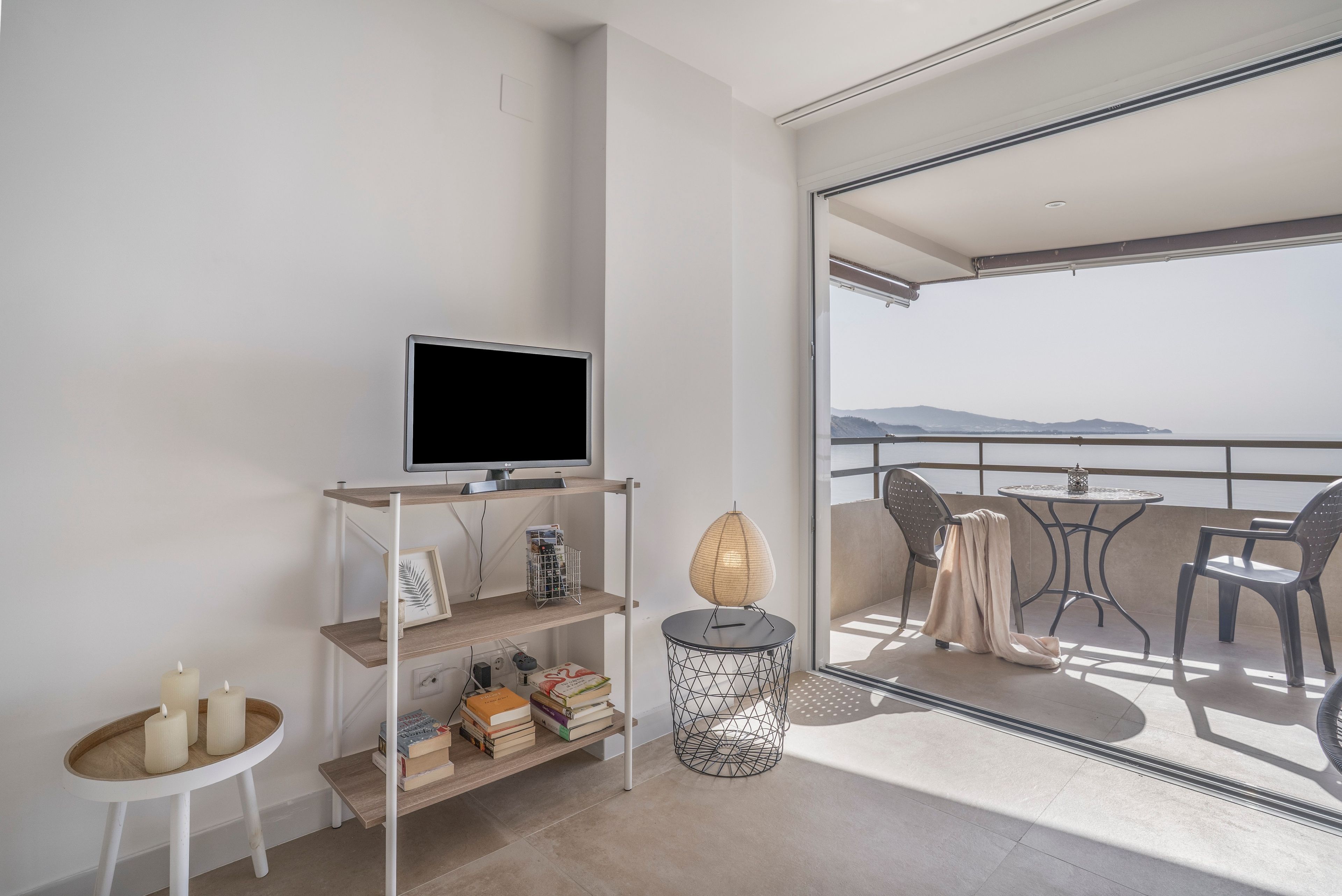 A modern living room with a flat-screen TV, books, a small table with candles, and a view of the sea through a balcony.
